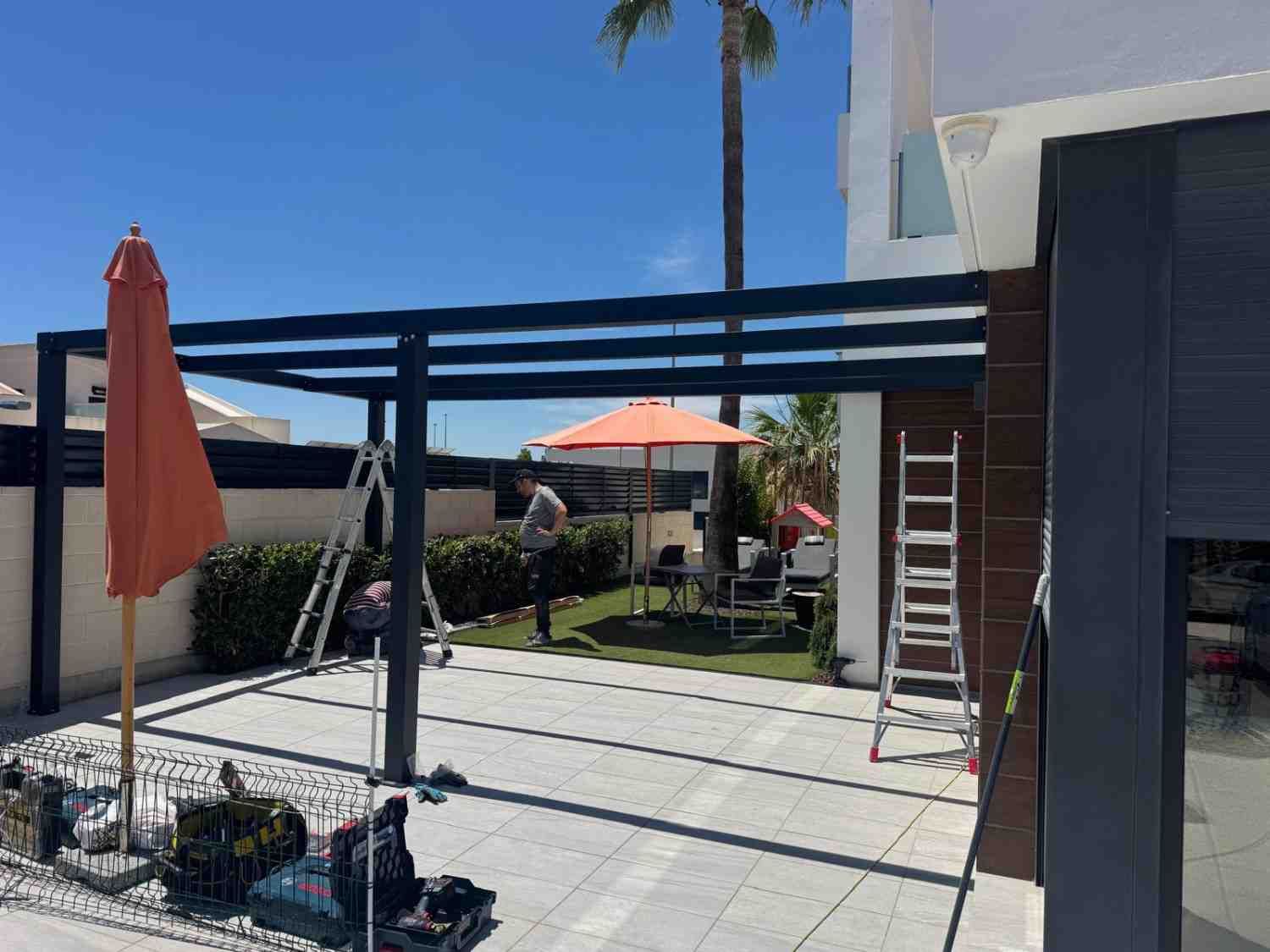 Patio con sombrillas naranjas, sillas de jardín y una palmera junto a un edificio moderno bajo un cielo azul.