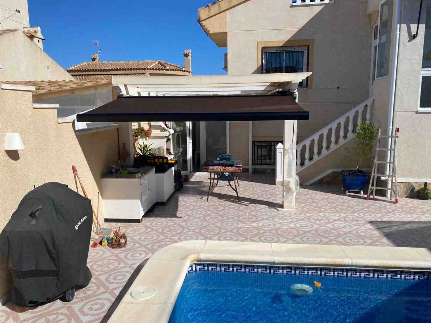 Patio trasero con piscina, zona de estar cubierta, barbacoa y cielo azul.