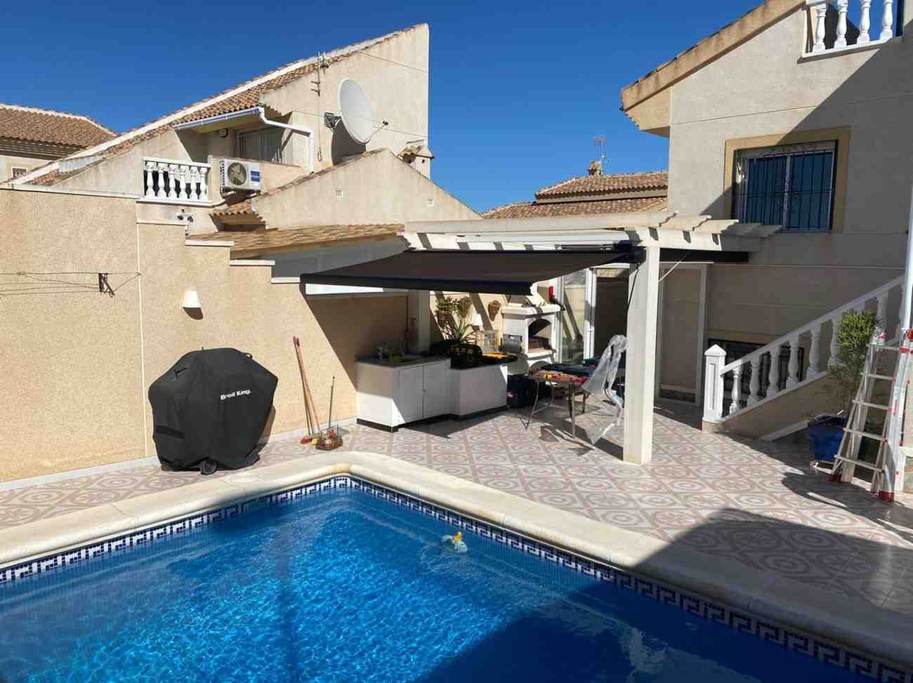 Patio trasero con piscina azul, paredes de estuco beige y una zona de bar exterior a la sombra.