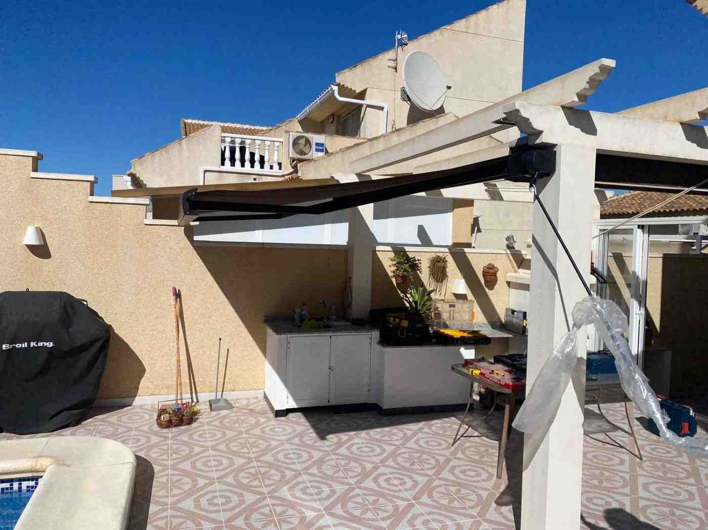 Patio con toldo retráctil beige, parrilla para barbacoa y plantas en macetas bajo un cielo azul brillante.