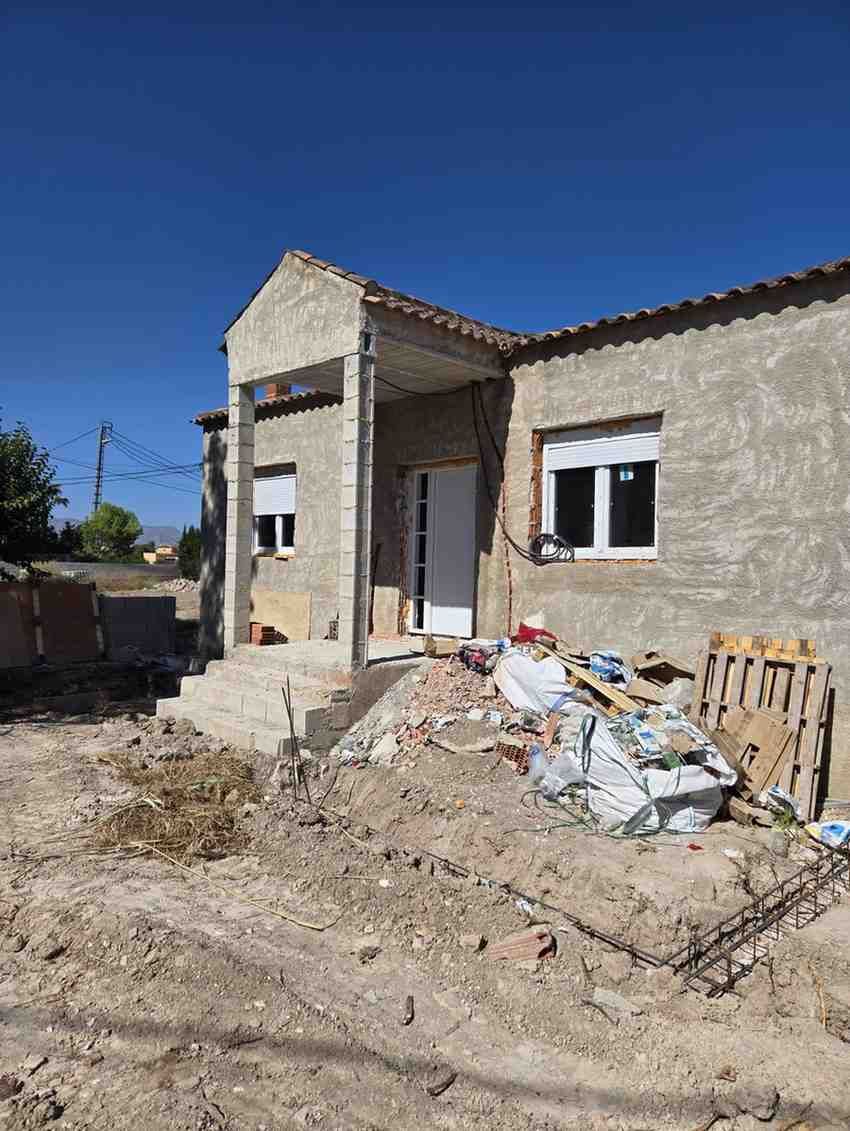 Casa de piedra parcialmente construida con escombros y restos de construcción en el exterior bajo un cielo azul despejado.