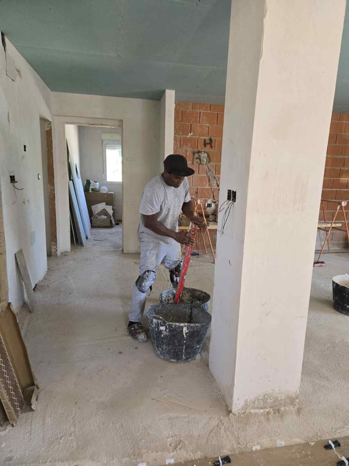 Un trabajador pinta una columna dentro de una habitación sin terminar, con herramientas y cubos en el suelo.