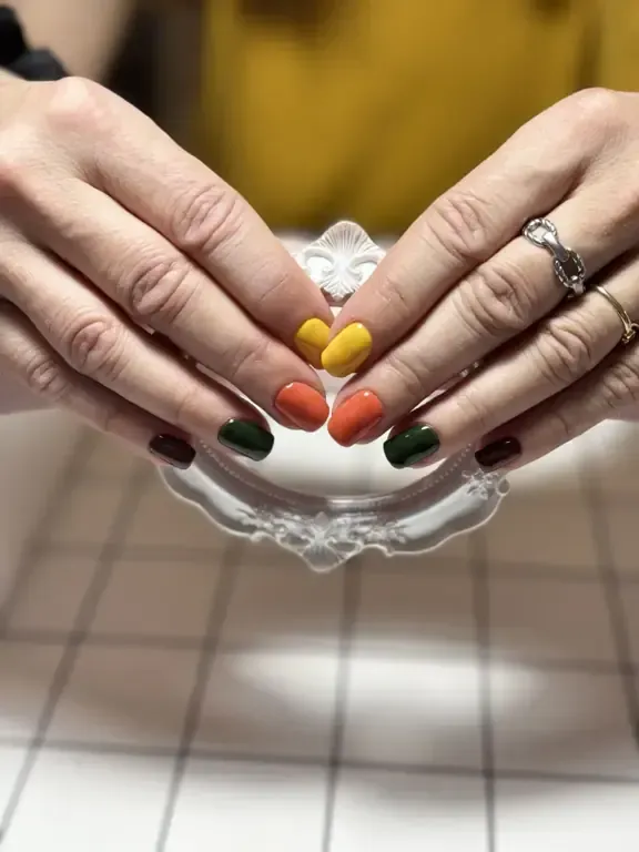 Manos con uñas ovaladas pintadas de amarillo, naranja, verde y burdeos, apoyadas sobre una superficie blanca decorativa.