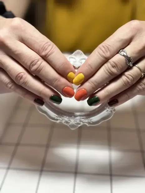 Manos con uñas ovaladas pintadas de amarillo, naranja, verde y burdeos, apoyadas sobre una superficie blanca decorativa.