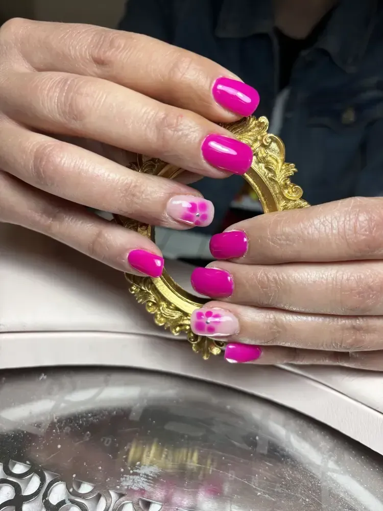 Manos con esmalte de uñas rosa brillante, sujetando un marco dorado decorativo. Dos uñas tienen un diseño rosa y blanco.