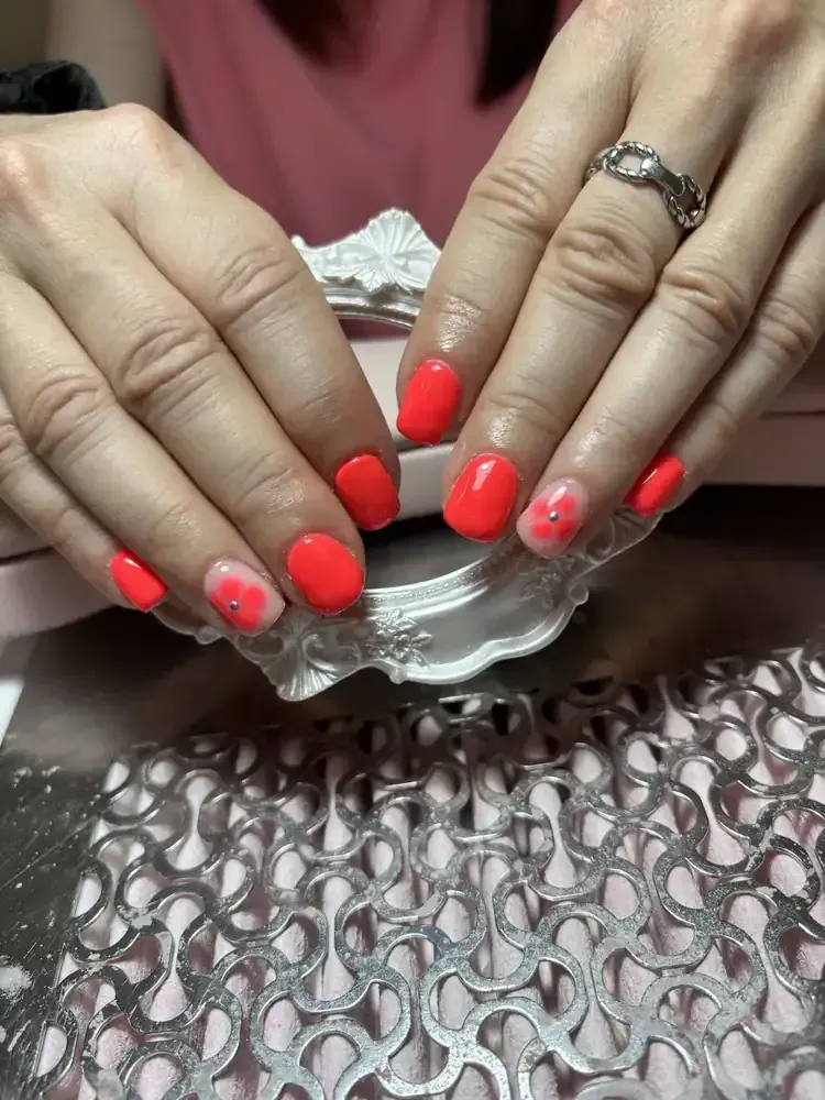 Manos con uñas de color naranja brillante, una de ellas con pequeñas joyas. Fondo rosa con detalles metálicos ornamentados.