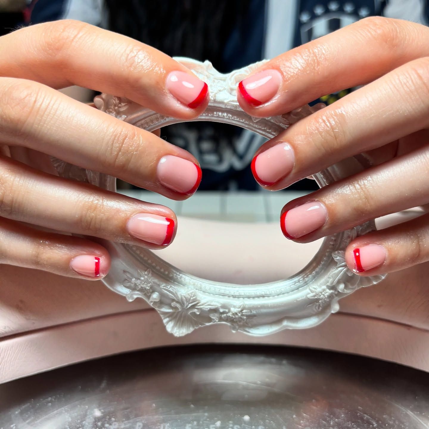 Manos con uñas con puntas rojas que sostienen un marco blanco adornado sobre una superficie rosa.