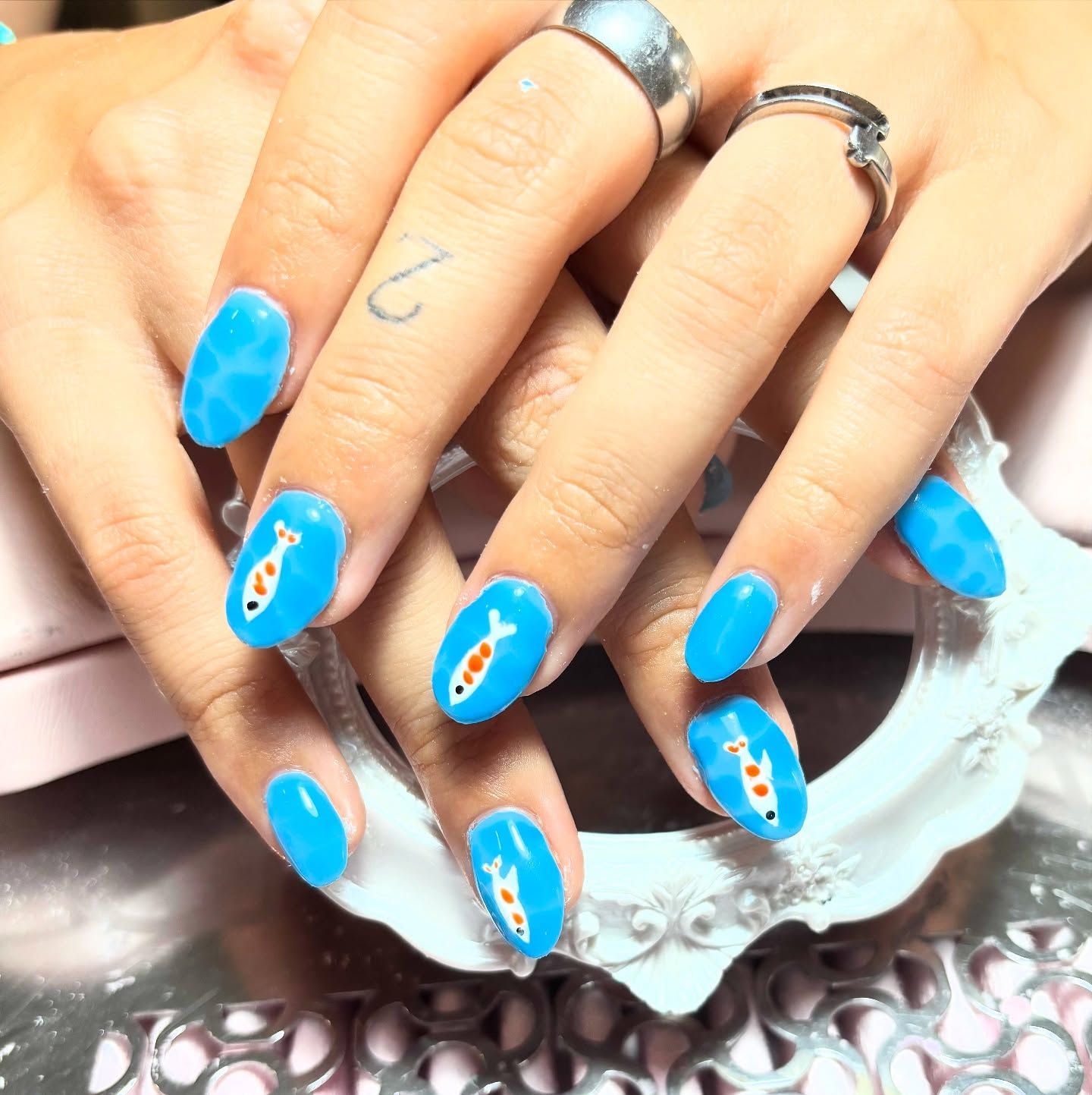 Uñas de color azul brillante con diseño de peces koi, mano con anillos plateados y un pequeño tatuaje.