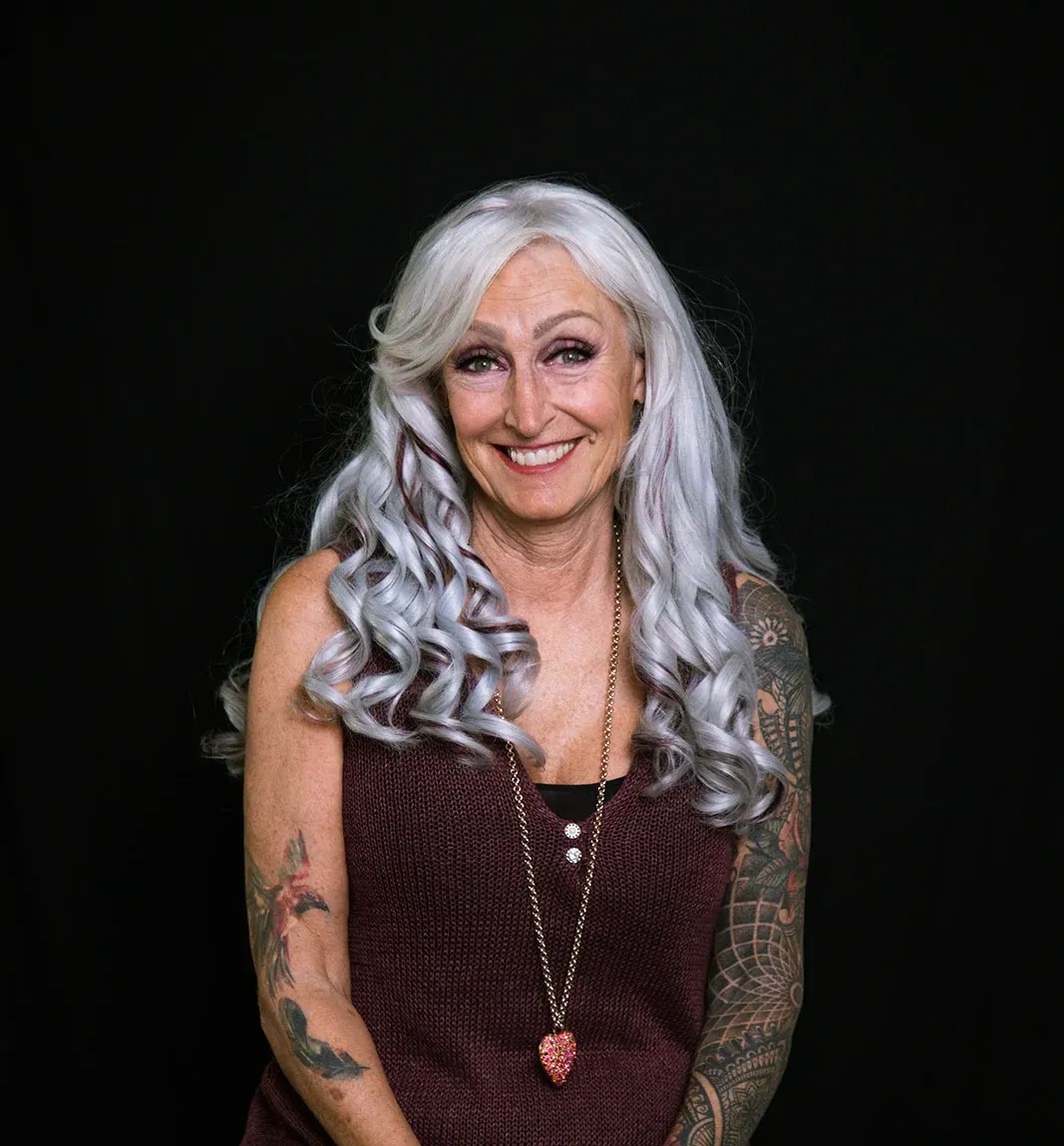 Una mujer con cabello gris y tatuajes en sus brazos está sonriendo.