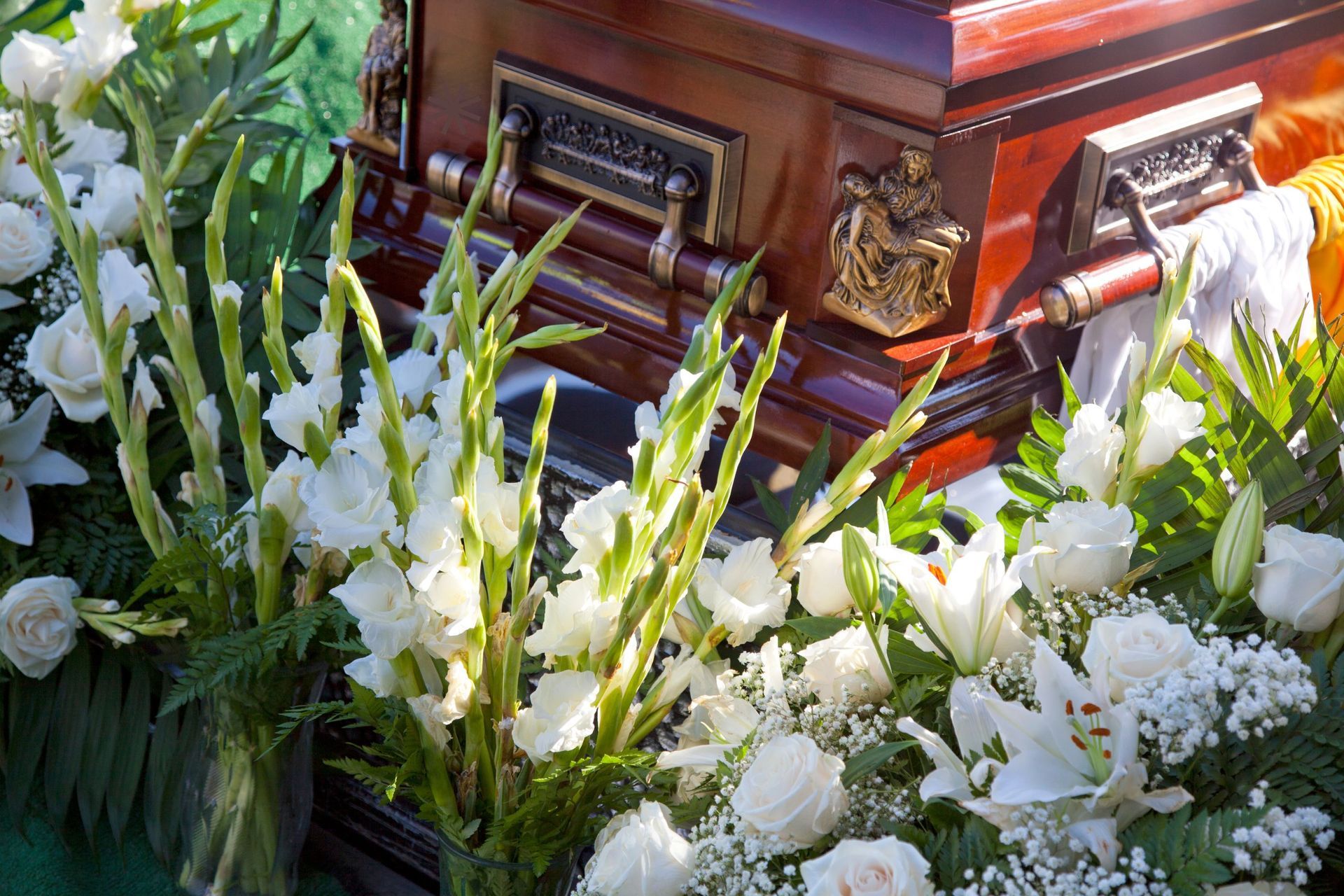 Weiße Trauerblumen neben einem polierten Holzsarg mit goldenen Griffen und einem Strauß roter Rosen.