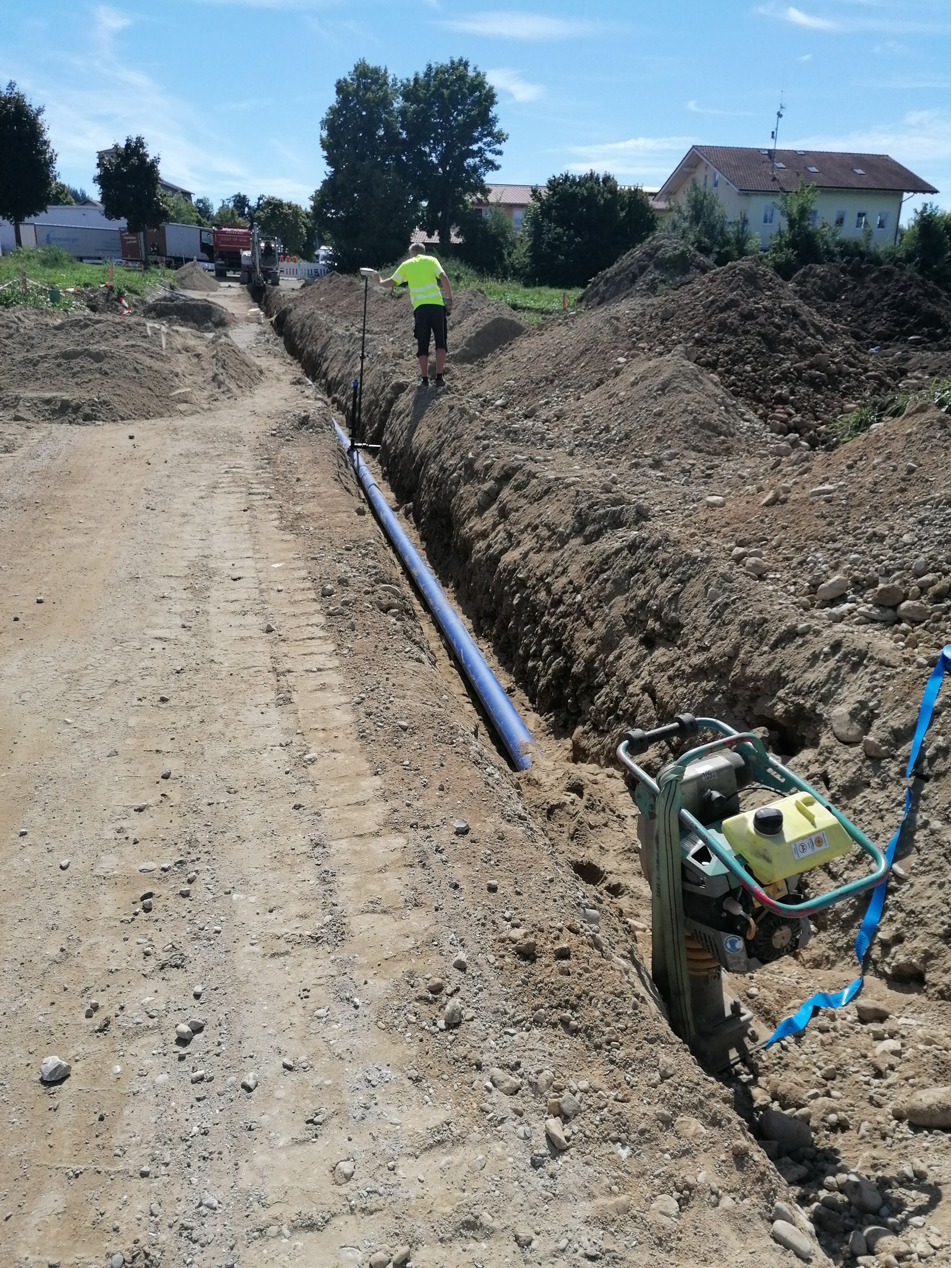 Bauarbeiter in gelber Warnweste steht in einem langen Graben mit verlegtem blauen Rohr, neben dem eine Rüttelplatte steht.