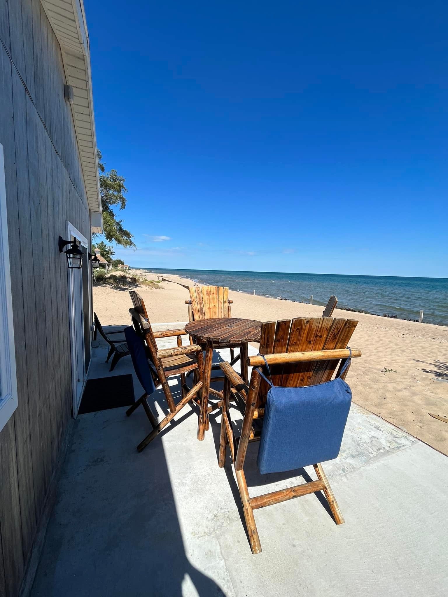 A rustic wooden patio set sits on a concrete slab next to a building, overlooking a sandy beach and blue ocean.