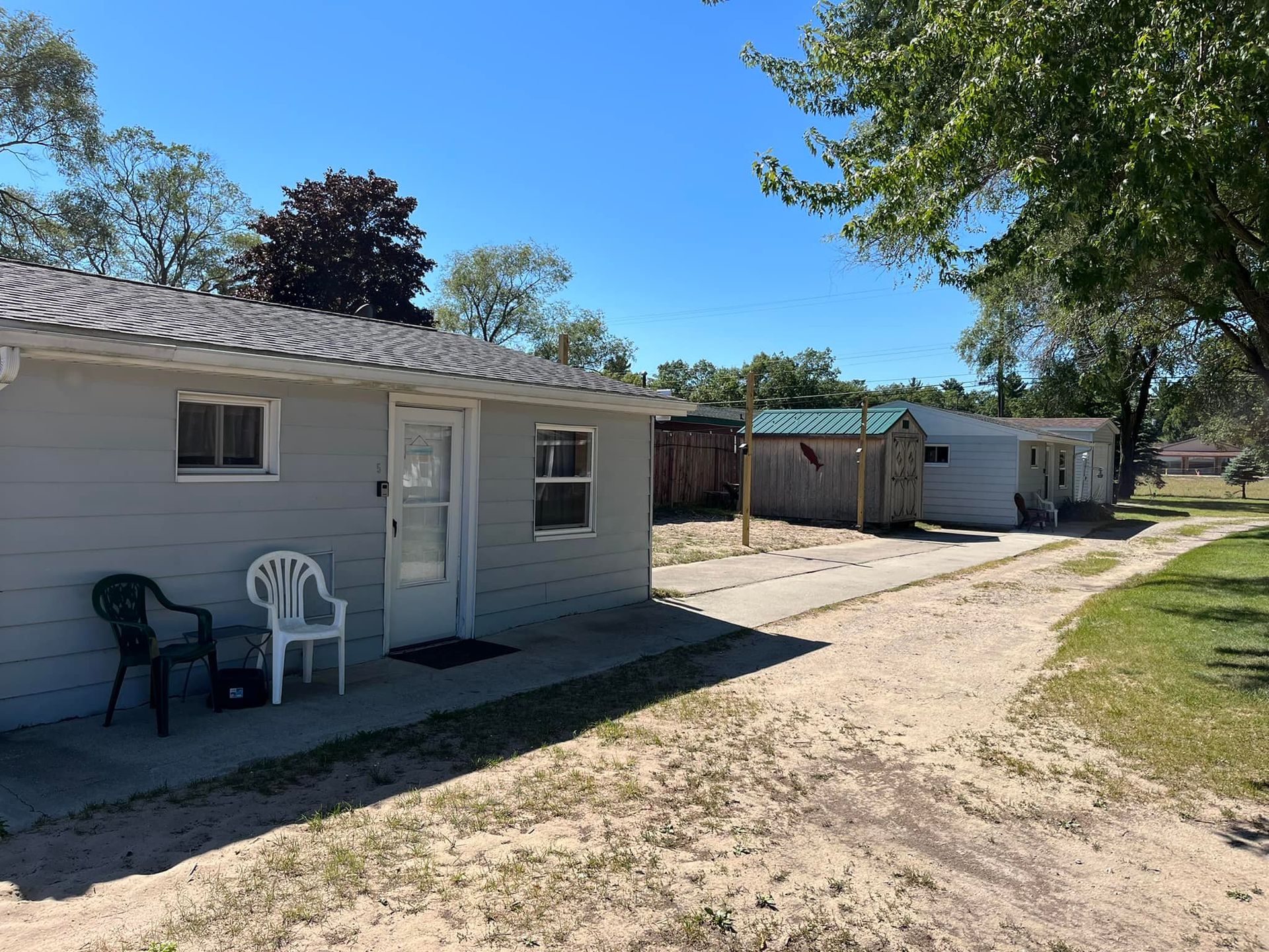 Light gray cottage with two chairs on a sandy path, leading to neighboring cabins under a clear blue sky.