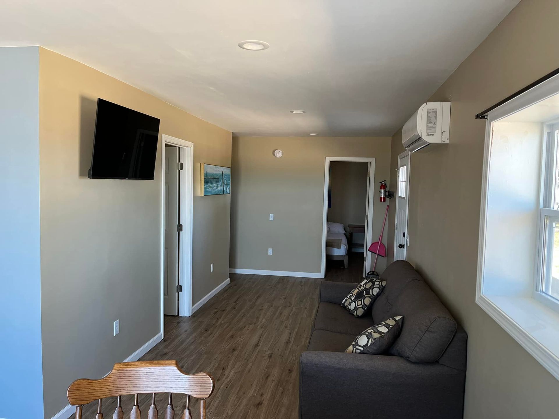 A living room with light brown walls, a grey sofa, a wall-mounted TV, wood-look flooring, and an open doorway.