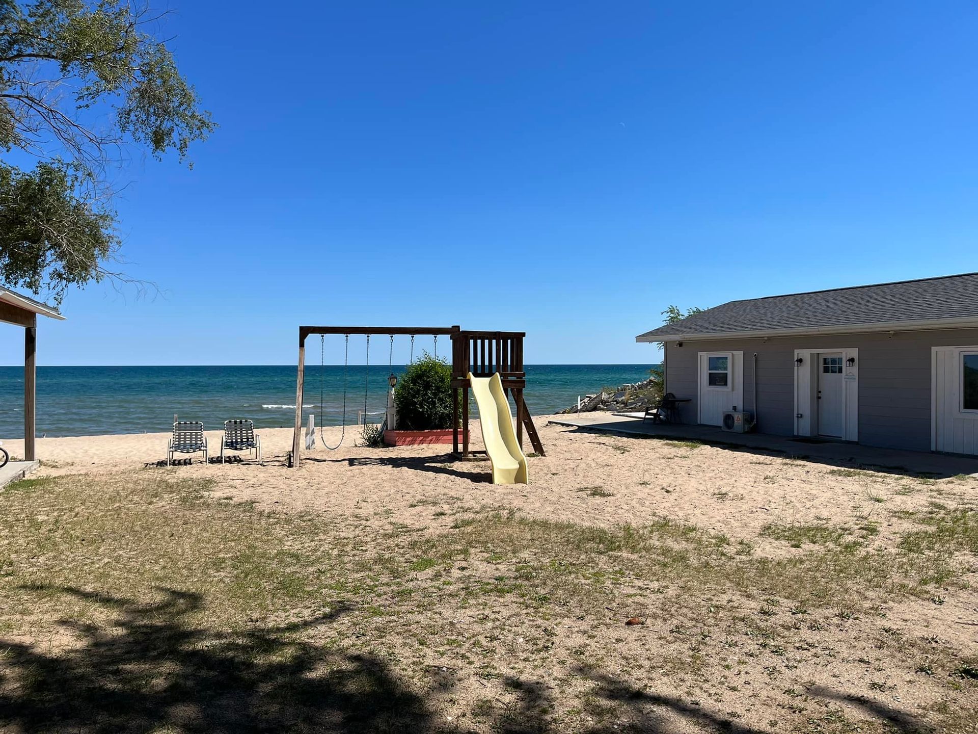 A sandy beach area with a swing set, yellow slide, and two chairs near a small building on a sunny day by the blue water.