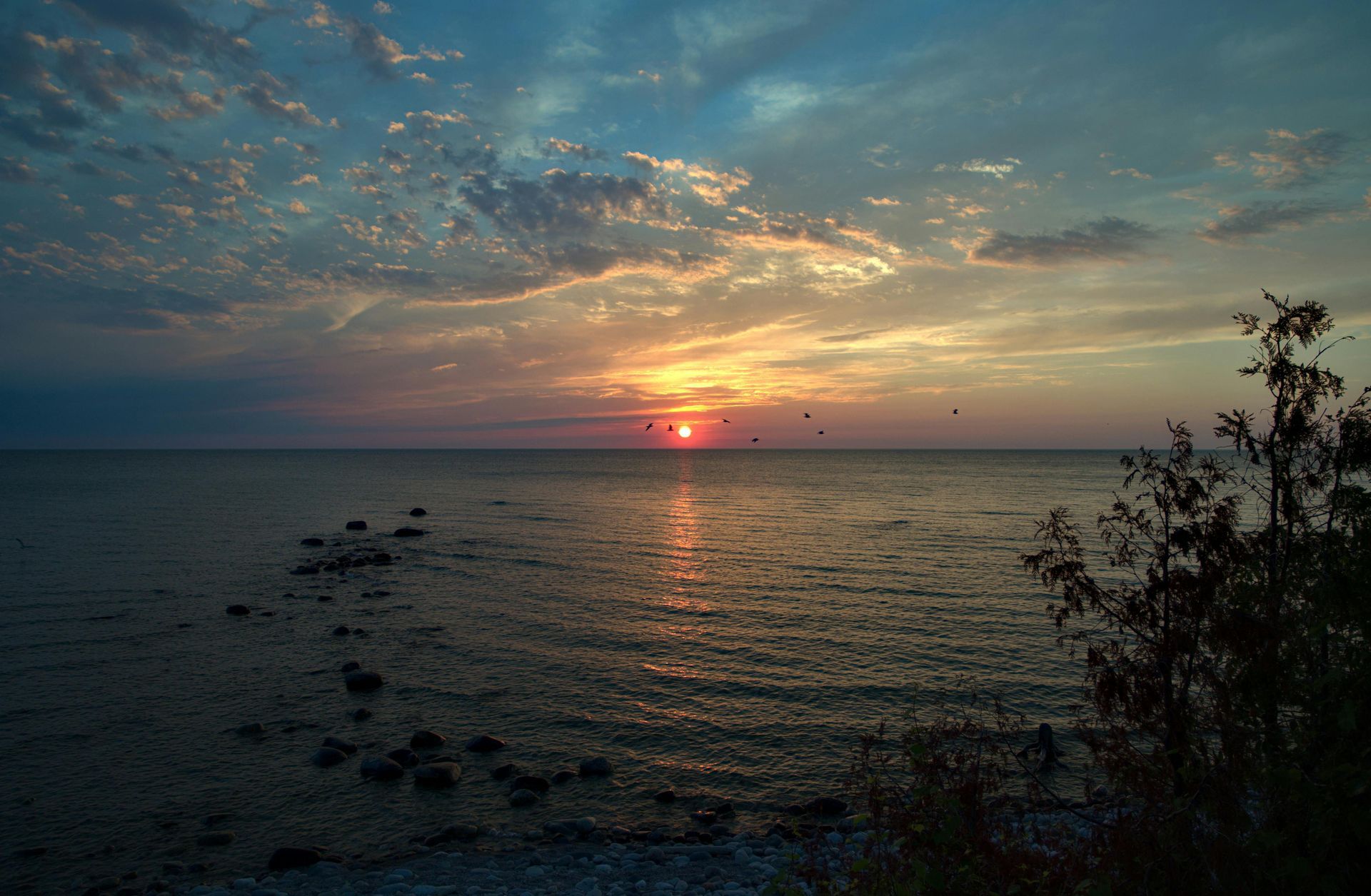 A sun sets over a calm ocean, casting a golden glow on the water with scattered clouds above and rocks near the shore.