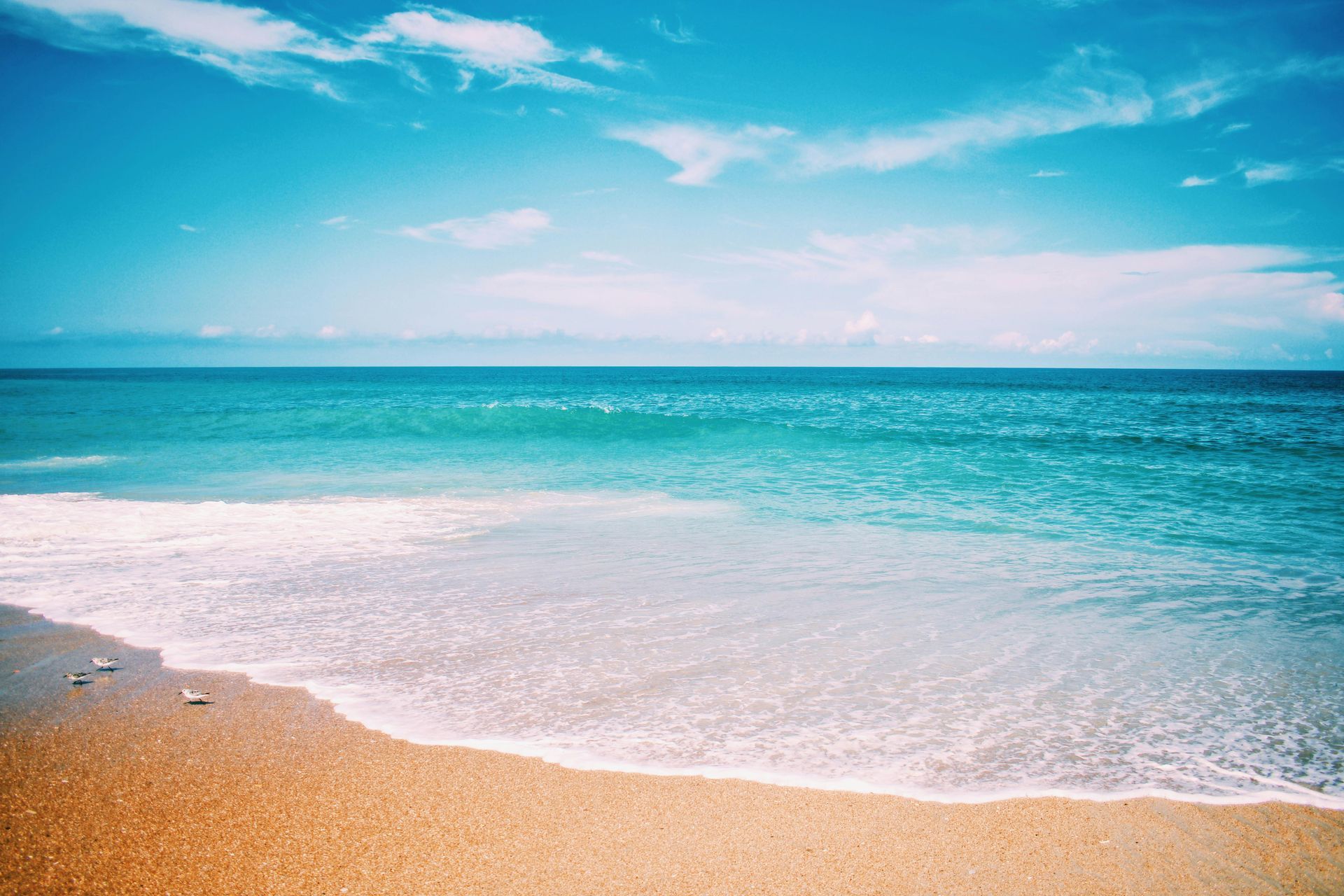 A sunny beach with golden sand meeting clear turquoise water under a vibrant blue sky with soft white clouds.