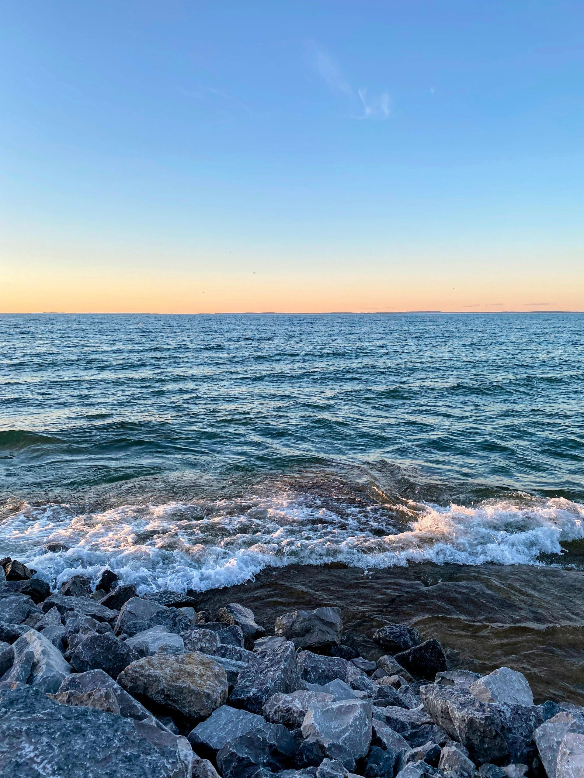Gentle waves roll toward a rocky shoreline under a serene, golden-hued sunset sky.