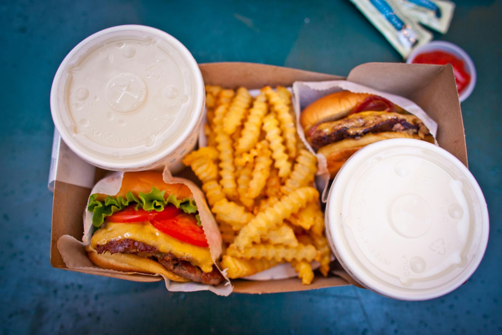 A cardboard takeout container holds two cheeseburgers, a large portion of crinkle-cut fries, and two drinks.