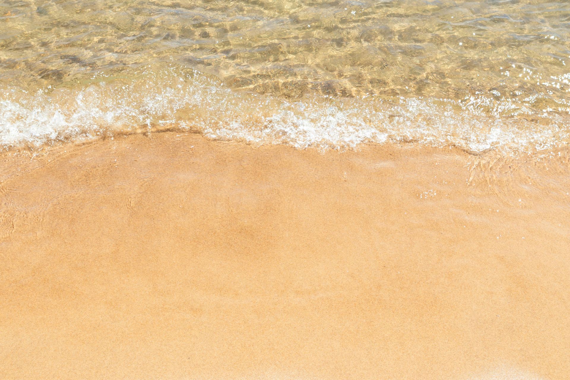 Clear water gently washing over light tan sand on a beach.