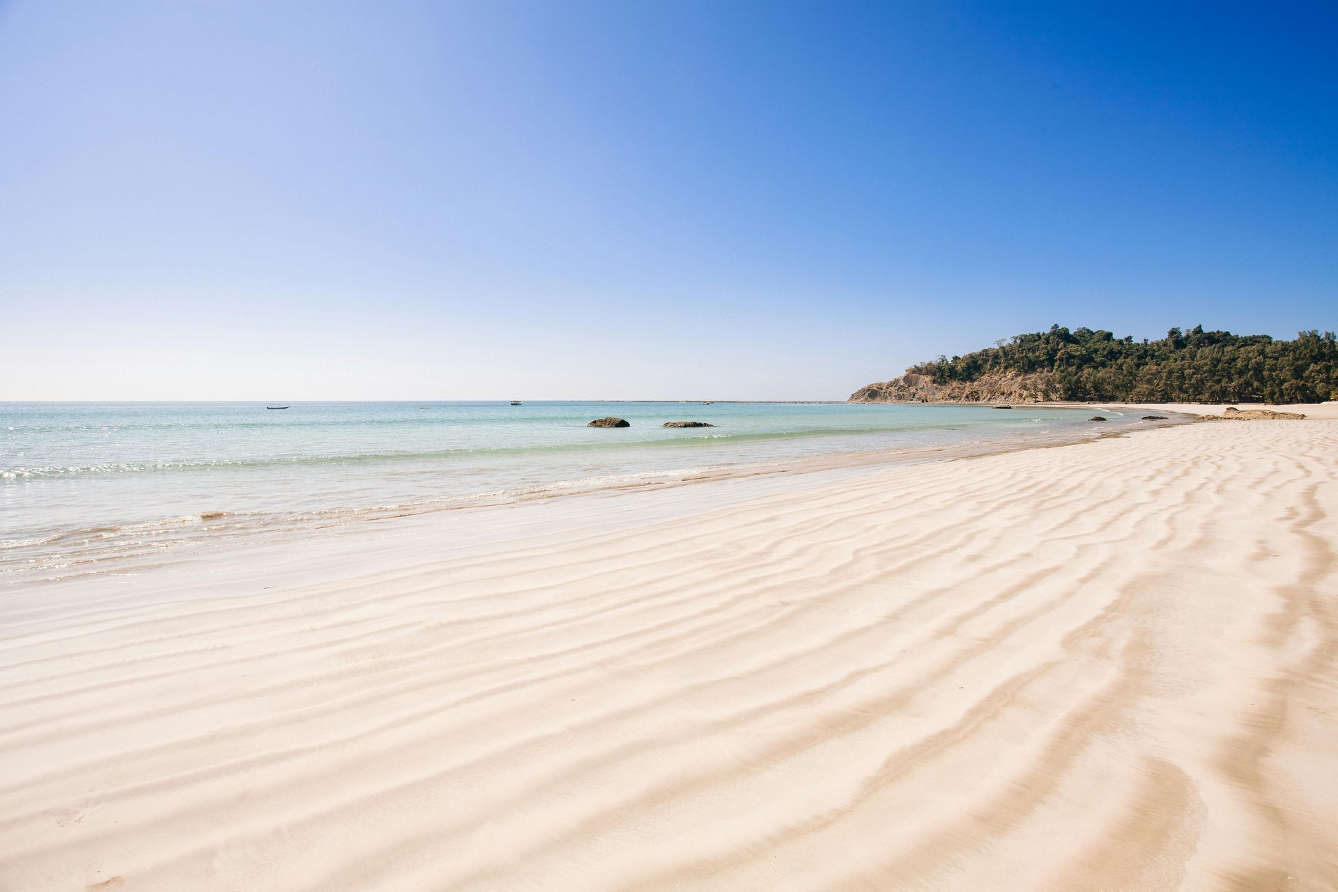 Rippled light sand leads to calm turquoise water under a clear blue sky, with a lush green headland in the distance.