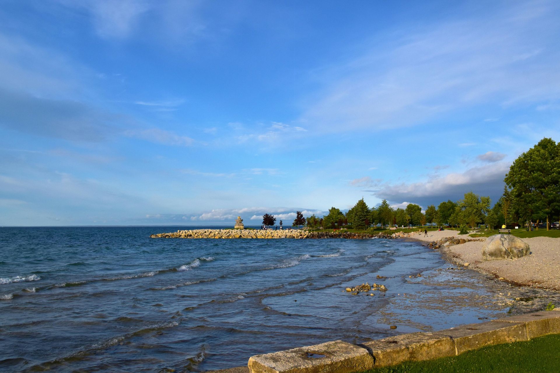 A rocky shoreline with small waves, trees, and a stone jetty under a blue, cloudy sky.
