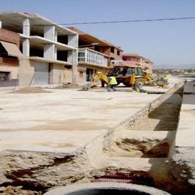 Obreros de la construcción manejan una retroexcavadora sobre una superficie de concreto frente a edificios de varias plantas en construcción.