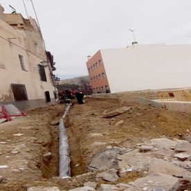 Una tubería yace dentro de una zanja abierta en una obra de construcción de tierra entre un edificio residencial y una estructura blanca moderna.