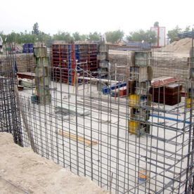 Una obra en construcción que muestra una valla de rejilla de barras de refuerzo en primer plano y estructuras de encofrado de hormigón al fondo.