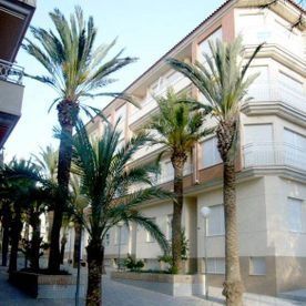 Un edificio de apartamentos de varias plantas con paredes de color beige y contraventanas cerradas, enmarcado por varias palmeras altas.