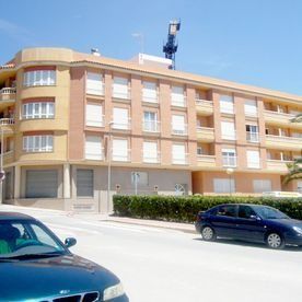 Un edificio residencial de cuatro plantas, de ladrillo color beige y con una esquina curva, visto desde una calle con coches aparcados.