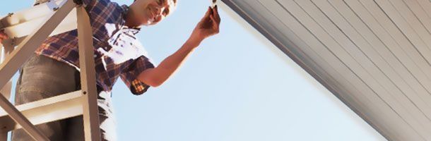 Una persona subida a una escalera extiende los brazos para pintar la parte inferior de un alero bajo un cielo azul brillante.