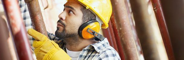 Un obrero de la construcción, con casco amarillo y protectores auditivos, levanta la vista mientras trabaja en tuberías metálicas.
