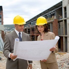 Dos personas vestidas con traje de negocios y cascos amarillos revisan un plano en una obra de construcción.