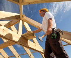 Un obrero de la construcción con casco sostiene un martillo mientras trabaja en la estructura de madera de un tejado bajo un cielo azul.