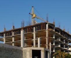 Un edificio de hormigón de varias plantas parcialmente construido, con barras de acero a la vista y una grúa amarilla contra un cielo azul despejado.