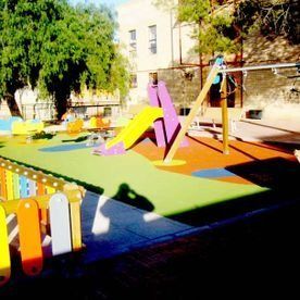 Un colorido parque infantil con un tobogán amarillo, suelo de goma verde y naranja, y una pequeña y animada zona vallada.