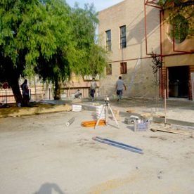 Una toma panorámica de una obra en construcción con tres trabajadores, un trípode y materiales de construcción cerca de un edificio de color beige.