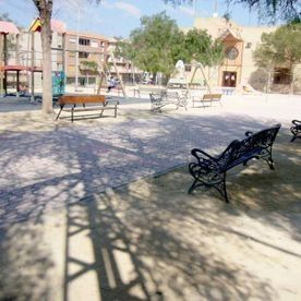 Un parque público soleado con un parque infantil, bancos, árboles que proyectan sombras y un edificio al fondo.