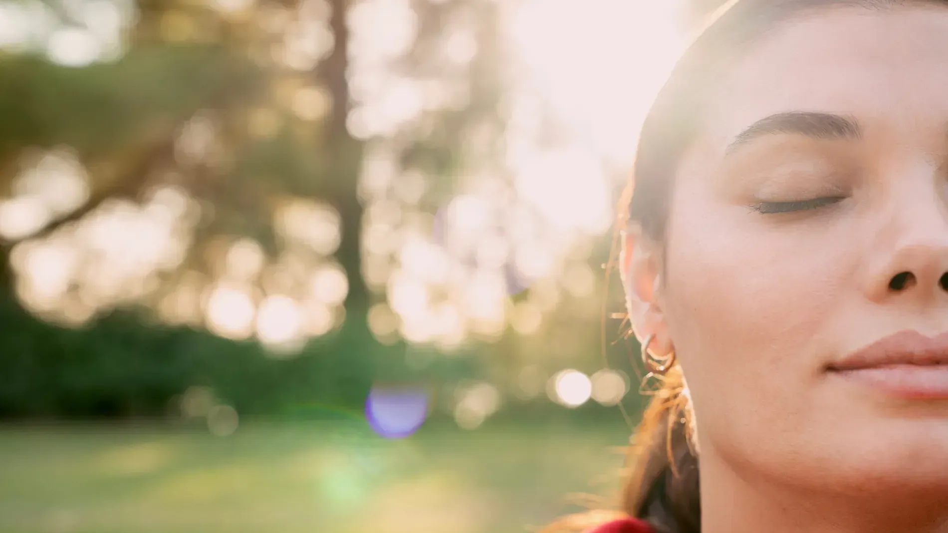 Primer plano de una mujer con los ojos cerrados bajo la cálida luz del sol, con un fondo de parque verde difuminado.