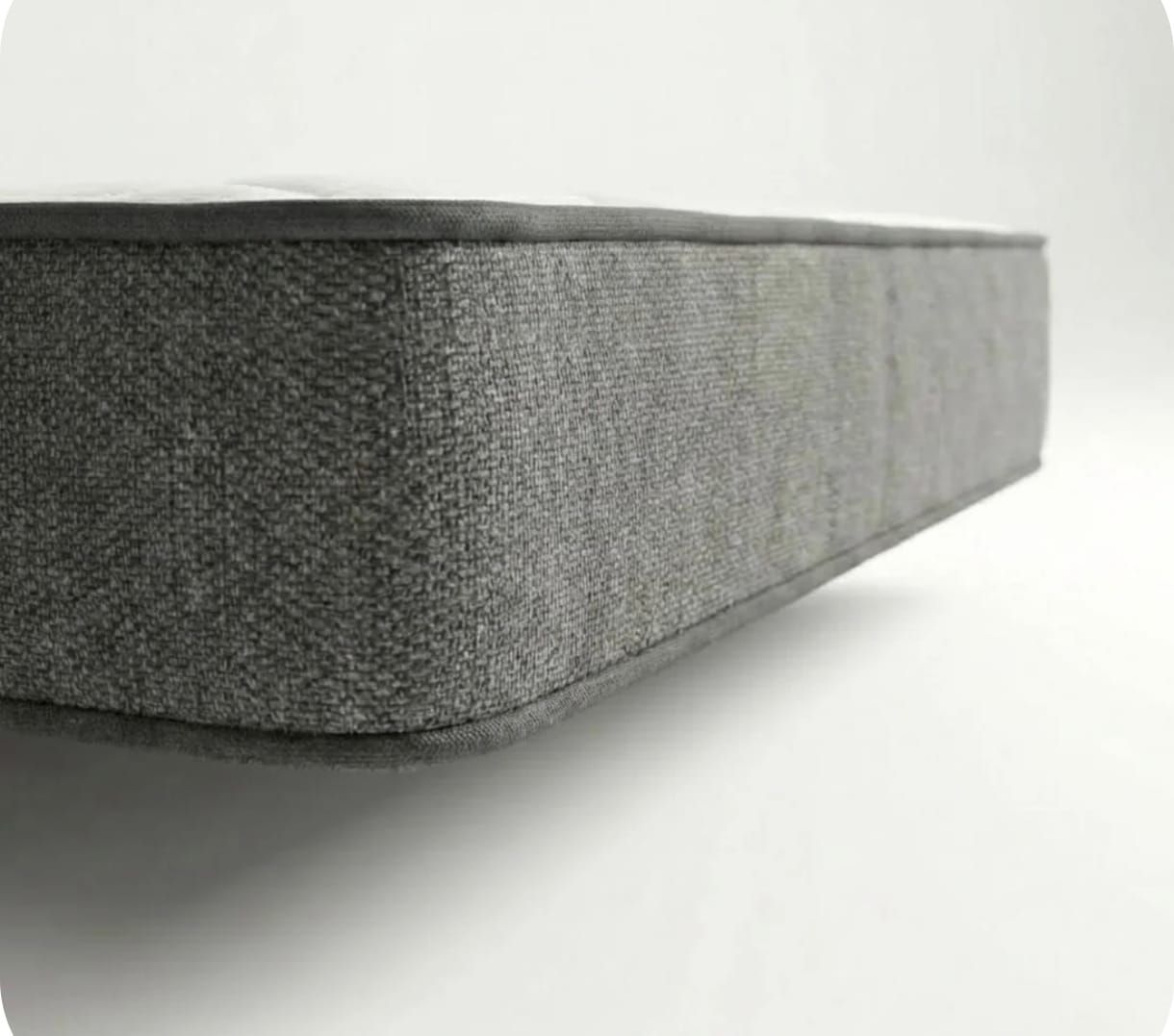 Primer plano del borde lateral de un colchón rectangular cubierto con una tela gris texturizada sobre un fondo blanco liso.
