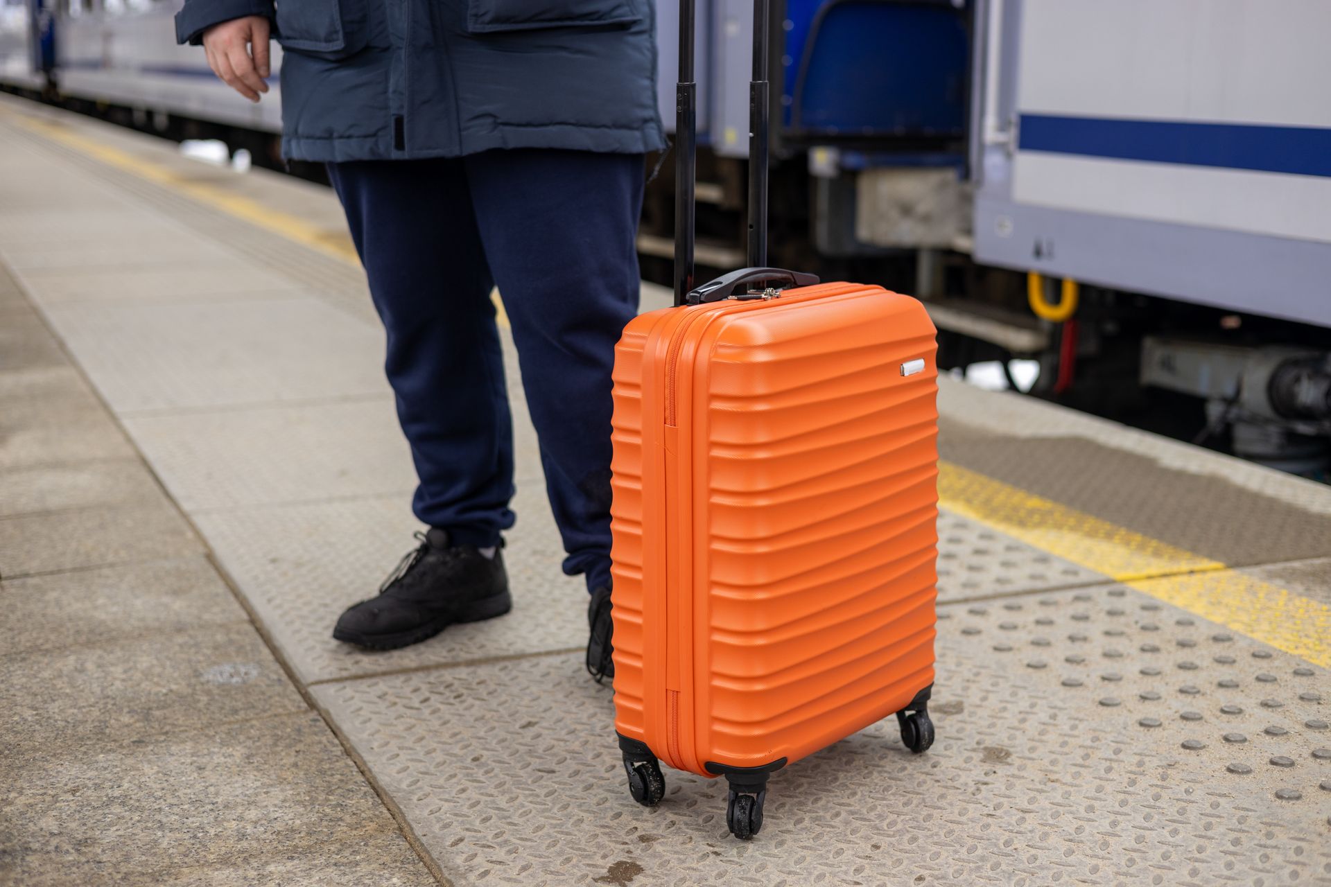 Une personne se tient sur le quai d'une gare à côté d'une valise à roulettes orange vif.