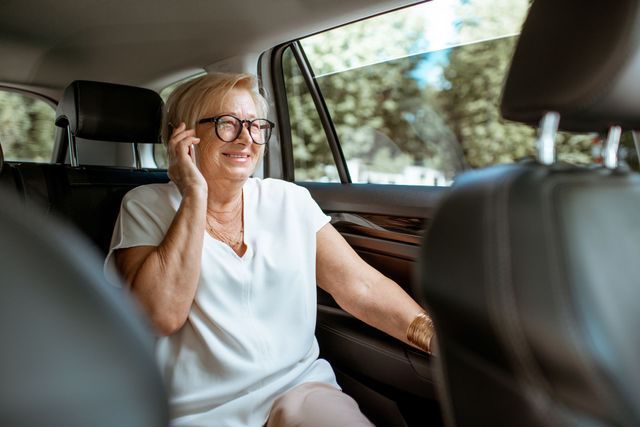 Une personne portant des lunettes est assise à l'arrière d'une voiture, souriante, un téléphone à l'oreille.