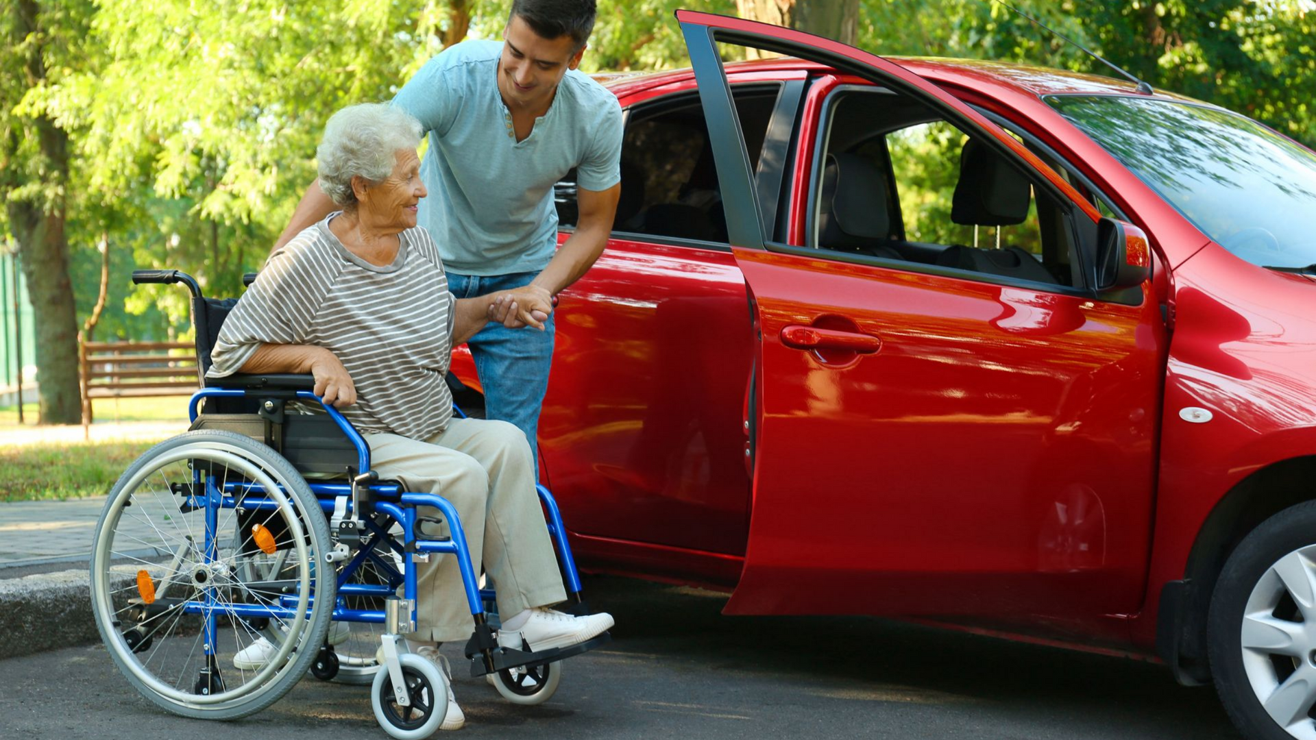Un homme aide une personne en fauteuil roulant à monter dans une voiture rouge garée dans une rue.