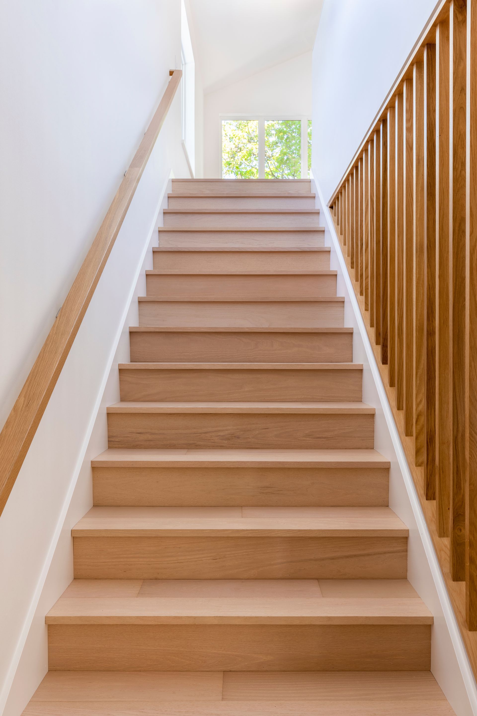 Escalier en bois clair avec des murs blancs et une rampe, menant à un palier baigné de lumière par une fenêtre