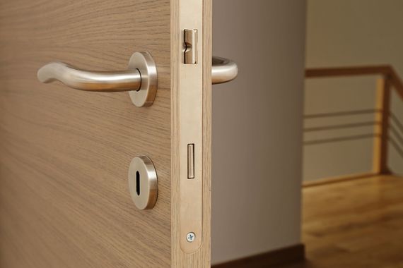 Gros plan sur une porte en bois avec une poignée et une serrure argentées, entrouverte dans un couloir.