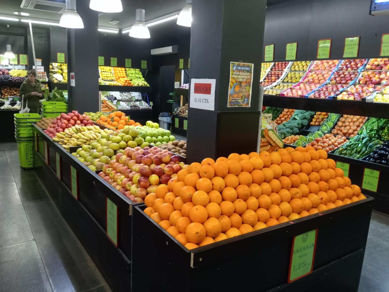 Una frutería de supermercado con naranjas, manzanas y otros productos en un interior oscuro.