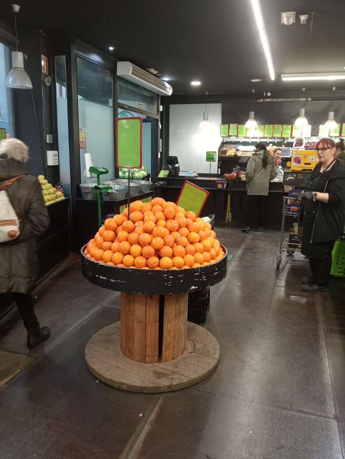 Una exhibición de naranjas en un carrete de madera en un supermercado. La gente compra, con un cajero tras el mostrador.