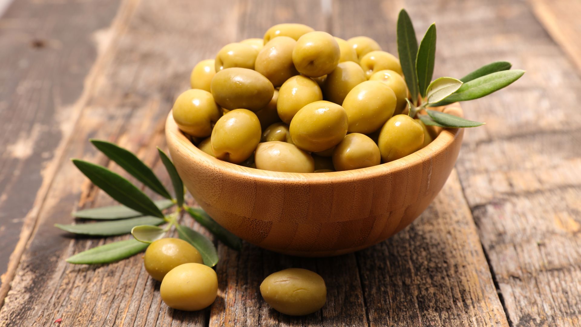 Un bol en bois rempli d'olives vertes est posé sur une table rustique, avec des brins de verdure et quelques olives éparpillées à proximité.