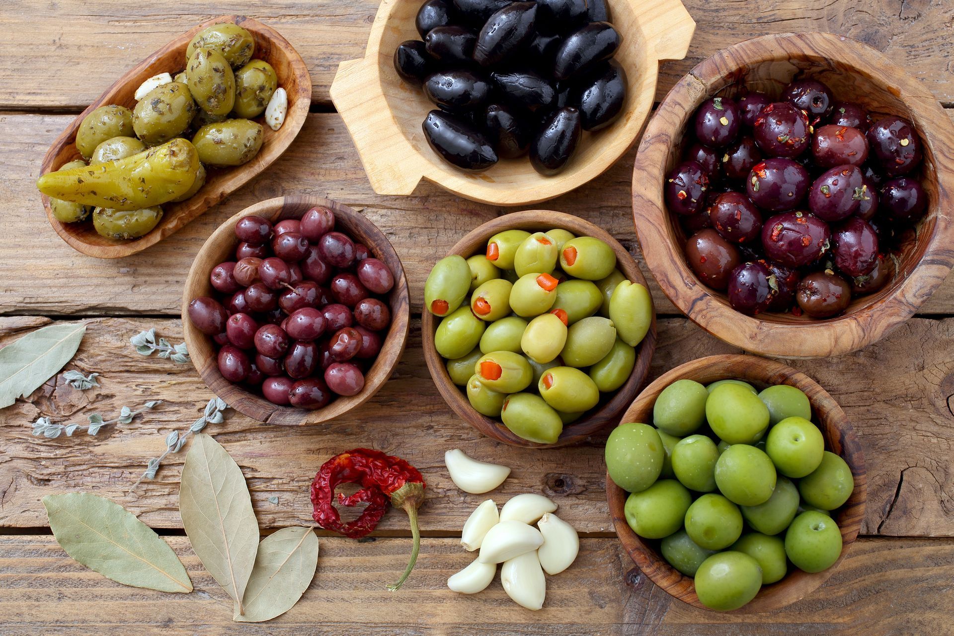 Olives et gousses d'ail assorties dans des bols en bois sur une table rustique, avec des feuilles de laurier et des piments rouges.